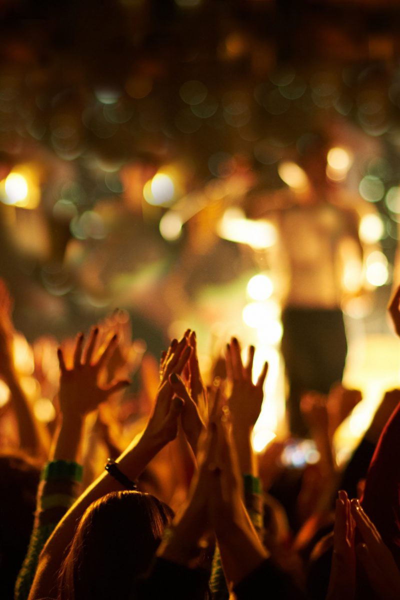 Audience with hands raised at a music festival and lights streaming down from above the stage. Selective focus.